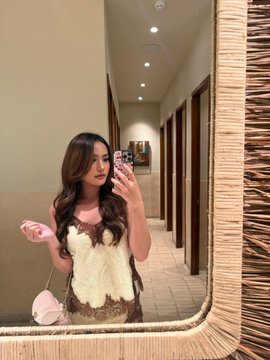 A mirror selfie of a woman with long brown hair wearing a brown lace top with a white furry bust detail, holding a pink phone, standing in a hallway with a textured mirror frame.