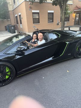 Two people sitting inside a black Lamborghini with bright green accents parked on a residential street, with a finger partially visible in the foreground.