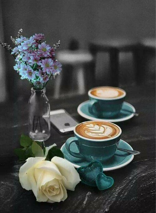 Two cappuccinos in teal cups sit on a table alongside a vase of blue and purple flowers, a white rose, and a smartphone.