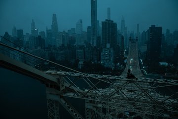 A moody, dark-toned view of a suspension bridge leading into a city skyline with tall skyscrapers.