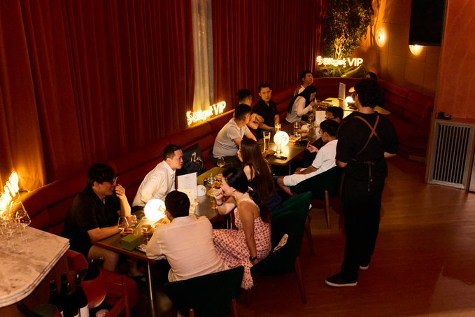 A group of people enjoying drinks and conversation at a dimly lit, intimate bar or lounge with red curtains and neon 'Subrise VIP' signs on the walls.