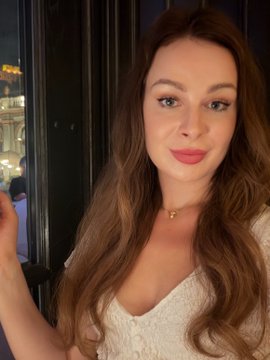 A close-up selfie of a woman with long wavy brown hair, wearing a white textured top and a delicate gold necklace, with blurred lights in the background.