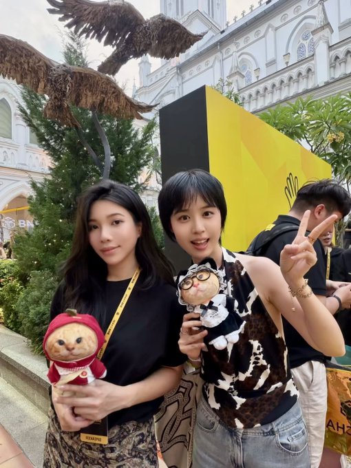 Two young women pose with plush toys in front of a Christmas tree featuring a large eagle decoration and a yellow "BNB" (Binance) sign, with the woman on the right flashing a peace sign while holding a clock-themed plush. The post tags @covachong, @fourdotmemezh, and @hajimi_CTO_BNB, suggesting this is a BNB community or Binance event photo celebrating their meetup.