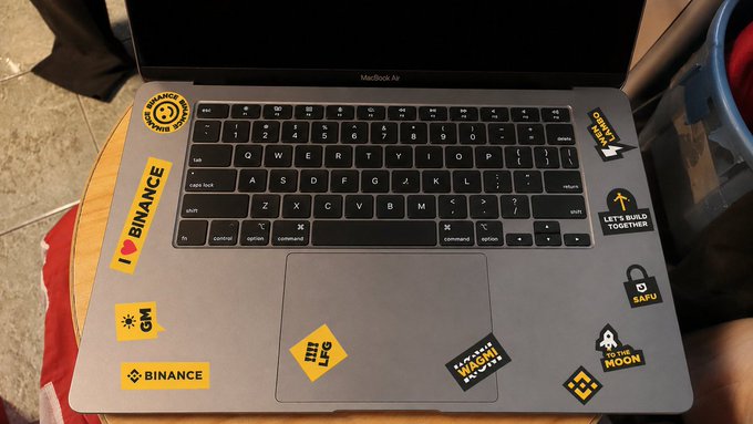 A MacBook Air laptop is decorated with various cryptocurrency-themed stickers, prominently featuring "BINANCE," "GM," "WAGMI," "TO THE MOON," and "SAFU" among others, with a yellow diamond-shaped "WTF" sticker near the trackpad. The stickers, which appear well-worn and likely accumulated over time, align with the caption's sentiment of being longtime companions from the beginning of a crypto journey.