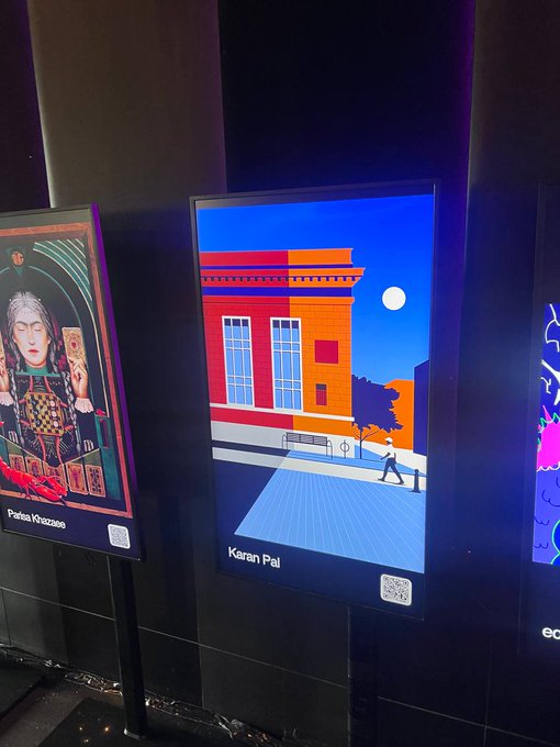 Two illuminated digital art displays in a dark setting, one labeled "Pietras Khozestoe" showing a portrait-style illustration and the other "Karan Pal" showing a stylized nighttime street scene with a red and orange building.