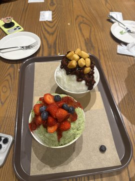 A tray with two bowls of shaved ice/dessert—one topped with fresh strawberries and blueberries on green (matcha) ice, the other with red beans and peanuts—on a wooden table.