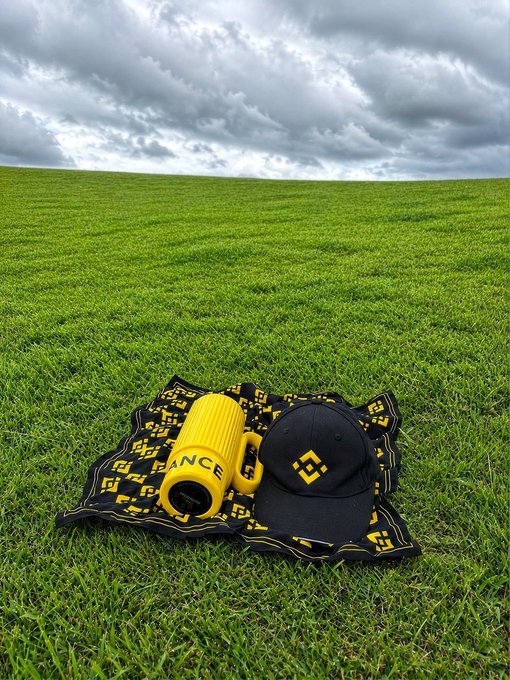 A yellow "BINANCE" branded water bottle and a black baseball cap featuring the Binance logo rest on a black and yellow Binance-patterned cloth, set against a vast green grassy field under a cloudy sky.