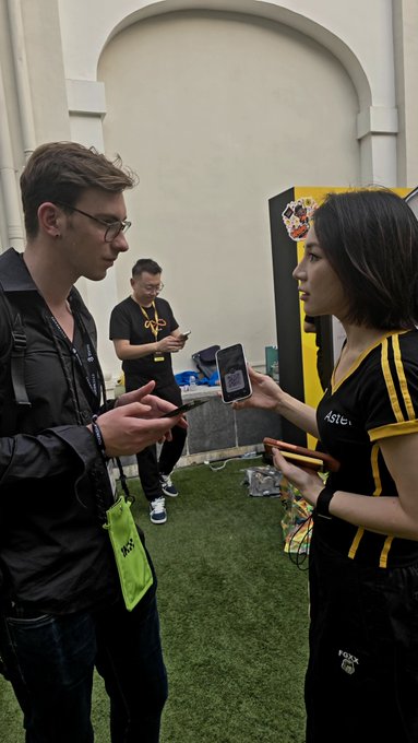 Two people, a man and a woman in branded yellow and black shirts, look at smartphones at what appears to be a gaming event, with another attendee in the background.
