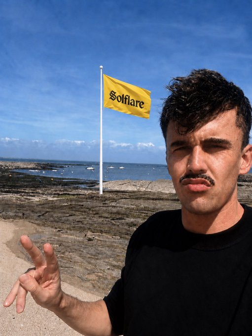 A man taking a selfie at a rocky beach making a peace sign, with a yellow Solflare flag in the background.