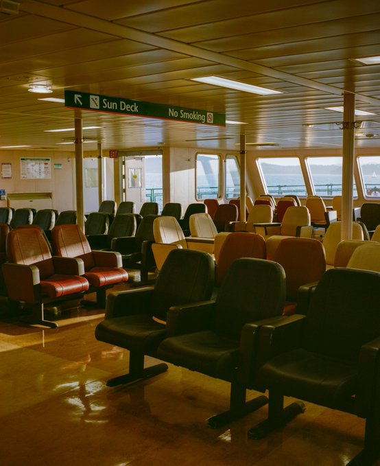 An empty ferry interior with rows of black, red, and yellow seats, featuring overhead signs reading 'Sun Deck' with an arrow and 'No Smoking'.
