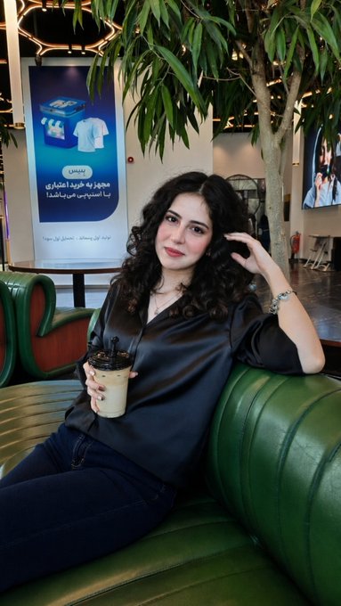 A young woman with curly dark hair sits on a green couch holding an iced coffee, with a digital ad banner in the background displaying Arabic text and the 'LAHAH' logo.