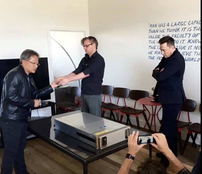 Three men in a room with a quote about knowledge on the wall; one man in a leather jacket hands an object to a man in glasses standing by a server-like machine on a table, while Elon Musk observes with arms crossed.