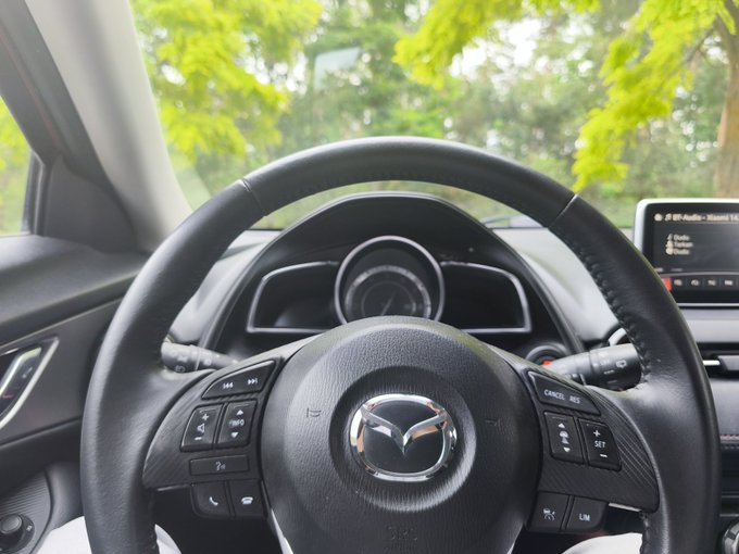 The image shows a car's steering wheel with a black color and a silver logo of a car brand. The perspective of the photo is from inside the vehicle, looking out at the surrounding area. There are trees visible outside the window, adding a natural element to the scene. The text in the image reads "I can' stand one week without @MarvinHarryP that's why its time to reunite today I am insanely hyped for the Solana summit, probably the best Event I've been so far Prepare yourself for some spamming, @SuperteamDE I am coming for you". The focus of the image is on the car and its surroundings.