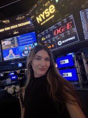 A woman with long dark hair posing in front of a wall of digital screens at the NYSE, with 'NYSE' and 'OG&E' logos visible on the displays behind her.