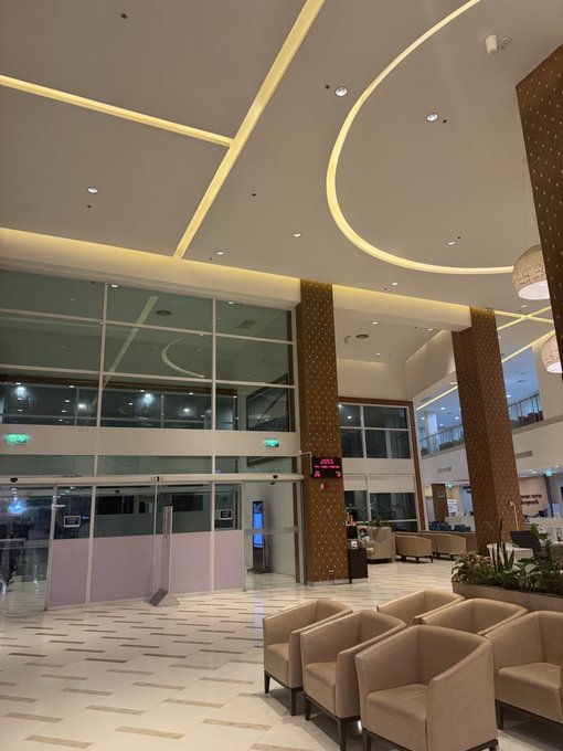 A dimly lit, modern hotel lobby with curved ceiling lighting and empty beige armchairs appears nearly deserted, with "2025" and "2024" digital display signs visible near the entrance doors. The image humorously suggests the caption's joke about crypto trading stress sending someone to seek refuge in an empty hospital or upscale lounge, though the surroundings look more akin to a luxury hotel than a medical facility.