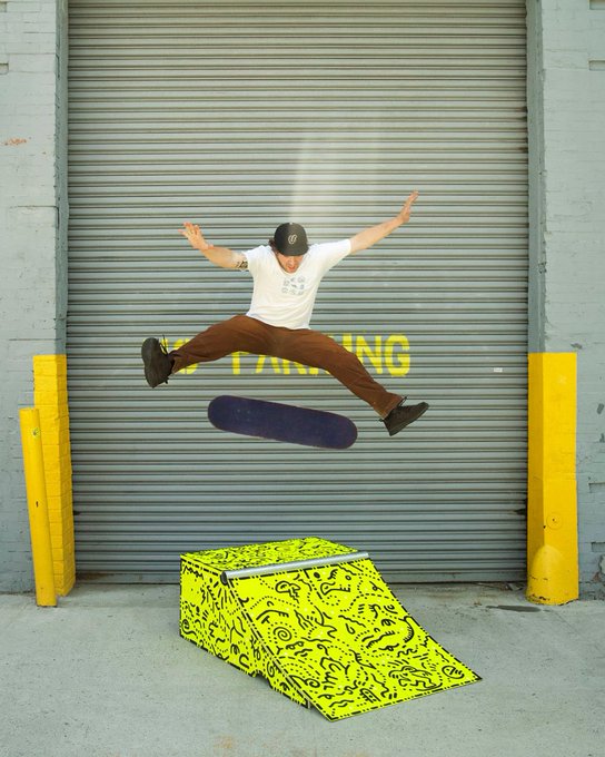A skateboarder mid-air performing a trick over a yellow ramp with doodle artwork, in front of a metal garage door marked 'NO PARKING'.