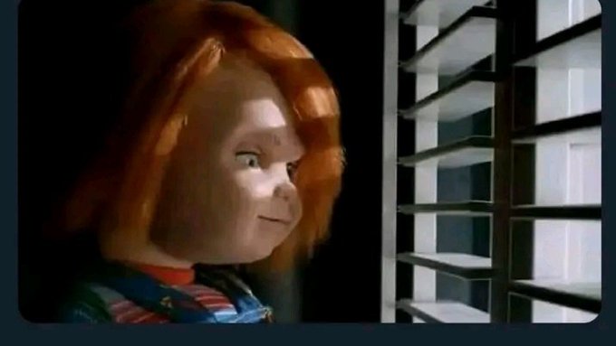 A scene from the Chucky horror franchise showing the iconic red-haired doll peering through white window blinds in dim lighting.
