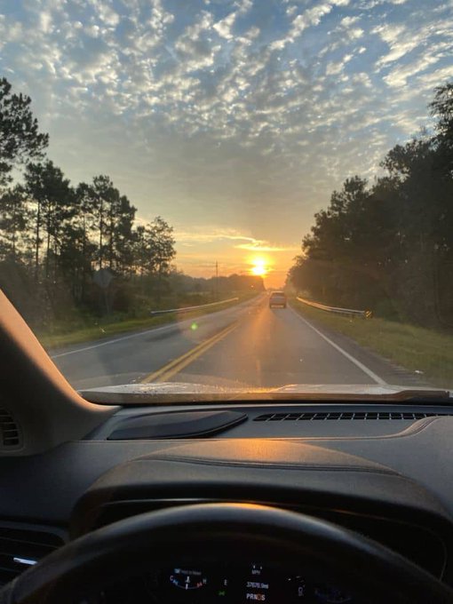 A scenic view from inside a car driving down a highway at sunset, with the dashboard and steering wheel visible in the foreground.