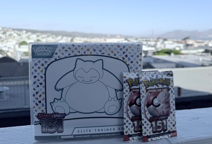 A Pokémon Scarlet & Violet 151 Elite Trainer Box featuring Snorlax sits on a railing with two 151 booster packs beside it, against a scenic hillside cityscape. The nostalgic 151-themed products connect to the post's excitement about a collector's giveaway or contest win tonight.