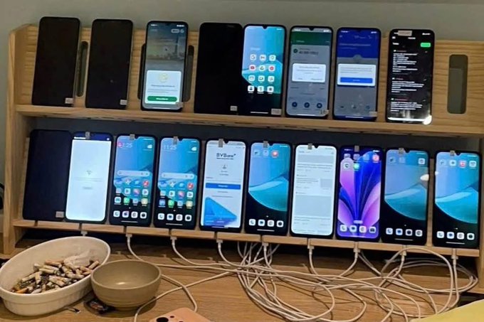A wooden display rack holding approximately 16 smartphones being tested or charged simultaneously, with cables running to each device and a small bowl of items nearby.