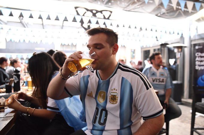 A young man wearing an Argentina #10 jersey drinks from a glass of beer at what appears to be a World Cup viewing event, with other fans and a Visa-sponsored shirt visible in the background.