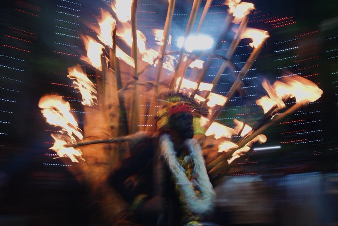 A blurred motion shot captures a performer adorned with a feathered headdress and surrounded by flaming torches, with a colorful light display creating streaks of color in the background. The image conveys the dynamic, cultural, and fiery energy referenced in the post about an abstract art piece celebrating traditional culture.