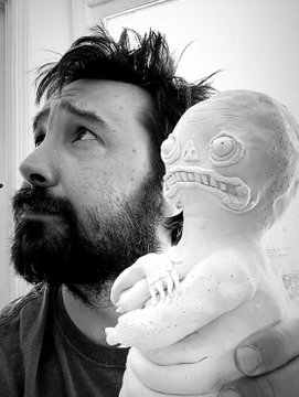 A black-and-white photo of a bearded man holding a small white statuette depicting a monkey holding a larger skull-like figure.