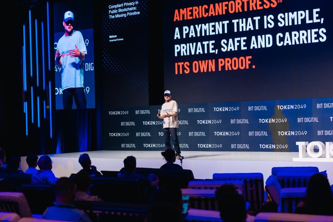 A presentation at TOKEN2049 showing a speaker on stage with a large screen displaying 'AMERICANFORTRESS — A PAYMENT THAT IS SIMPLE, PRIVATE, SAFE AND CARRIES ITS OWN PROOF.' under the title 'Compliant Privacy in Public Blockchains: The Missing Primitive'.