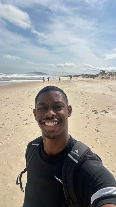 A selfie of a smiling young Black man wearing a black Reebok shirt and backpack on a sandy beach, with the ocean and distant figures visible behind him.