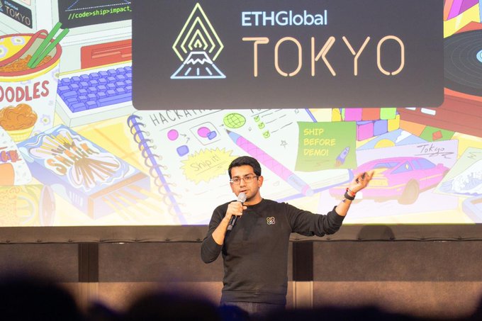 A man speaking into a microphone on stage at 'ETHGlobal TOKYO', gesturing toward a colorful backdrop displaying 'HACKATHON', 'SHIP BEFORE DEMO!', and other text.