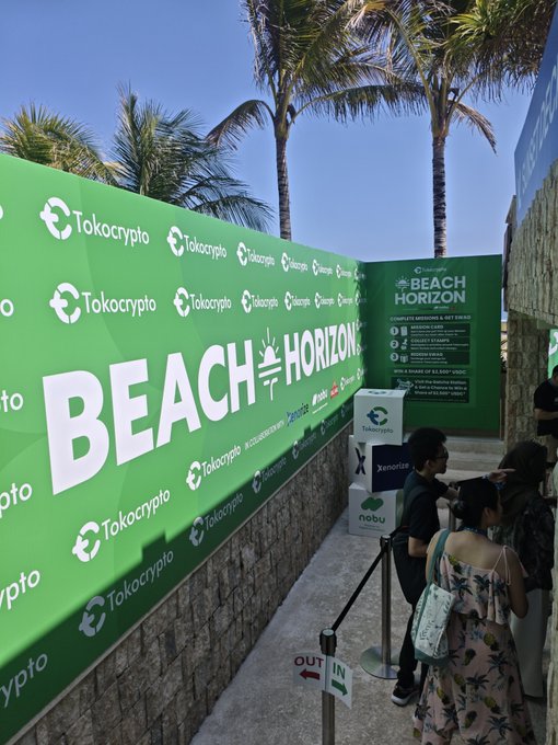Attendees gather at the entrance of the vibrant green "Beach Horizon" event hosted by Tokocrypto, featuring branded walls and a step-and-repeat backdrop under sunny palm trees. The image captures the welcoming atmosphere of the crypto gathering, with a queue forming near the "OUT" sign and informational displays about the event visible in the background.