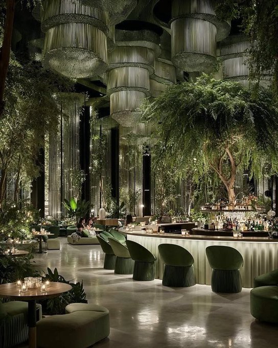 urns of light hang from the ceiling, casting a warm glow over a lush green interior with tables and chairs arranged for patrons to dine al fresco.