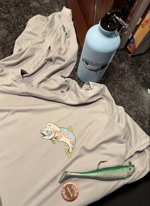 A light-colored hoodie featuring a colorful rainbow trout graphic is laid out alongside a matching enamel pin and a soft swimbait lure, with a blue "Water Hauler" branded water bottle in the background—showcasing fishing gear that complements the "#WHBTC faithful are thriving" caption, likely referencing the lifestyle brand's community enjoying outdoor activities.