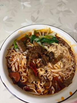A steaming bowl of Chinese-style braised beef noodles with bok choy and red peppers sits on a floral tablecloth. The image humorously contrasts the lighthearted food break with the accompanying frustrated post about being financially "screwed over" and pondering which cryptocurrency (BTC, ETH, SOL, or OKB) to invest in next.
