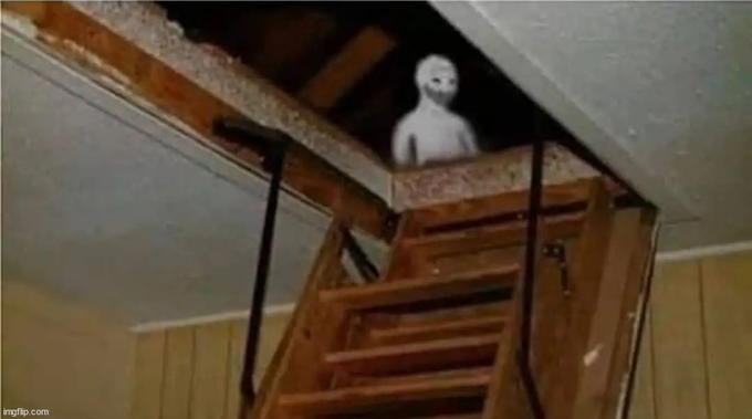 A creepy pale figure stands in the dark attic doorway at the top of a wooden staircase in a dimly lit house, creating a spooky scene.