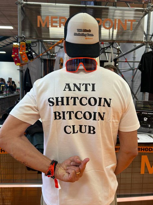 The image features a man wearing an anti-bitcoin t-shirt, sunglasses, and a hat with the words "anti-bitcoin club" written on it. The man appears to be making a peace sign with his hand while standing next to a table or counter. The background of the image is blurred, but there are some people present in the scene.

The post that the image is related to states that everything else is a scam except for Bitcoin. This message seems to be about Bitcoin and its supporters, possibly promoting their cause against Bitcoin.