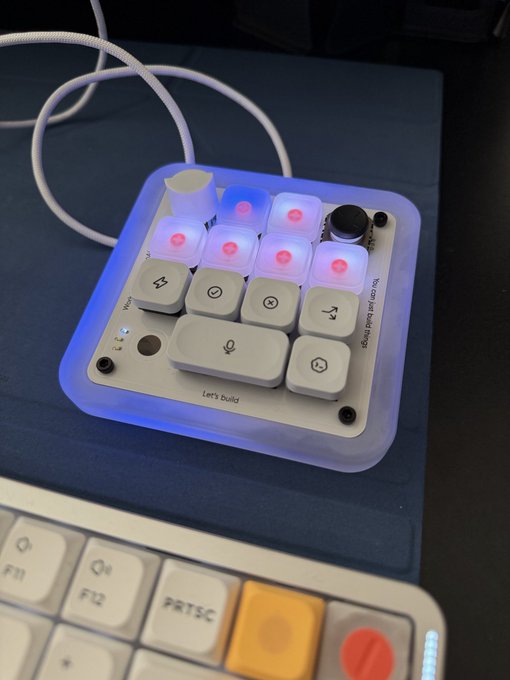 A small white macro pad with translucent backlit keys sits on a dark blue desk mat, reading "Let's build" and "Plugins just don't things" alongside a row of colorful keyboard keycaps (F11, F12, PRTSC) visible in the foreground—matching the poster's mix of excitement and buyer's remorse as they unbox their new programmable keypad.