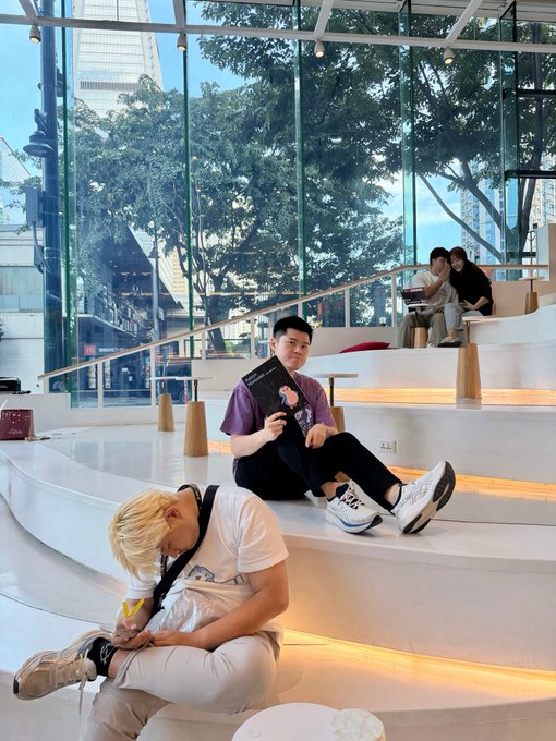Two young men sit on tiered steps in a modern, glass-walled building; one with blonde hair wears a white t-shirt and looks at his phone, while the other in a purple shirt holds a menu or pamphlet. In the background, two more people sit on higher steps, with a city street and trees visible through the large windows.