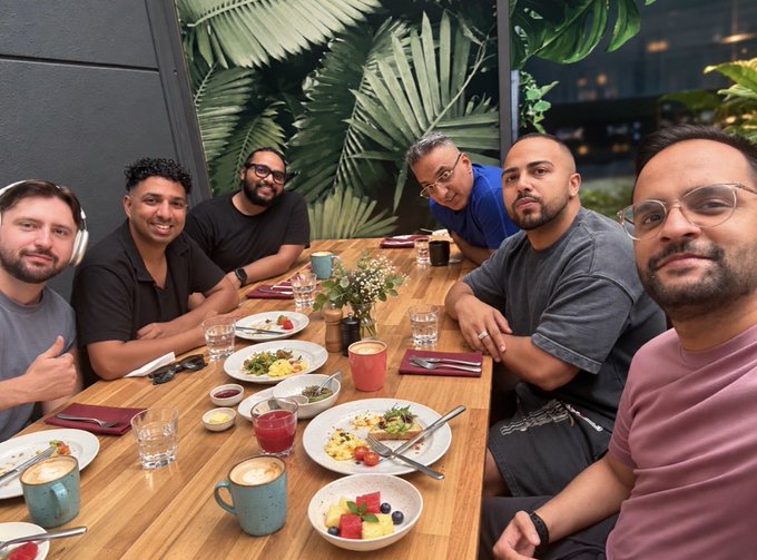 A group of six men smile for a selfie while seated around a wooden dining table featuring various plates of food, coffee cups, and tropical leaf decor in the background.