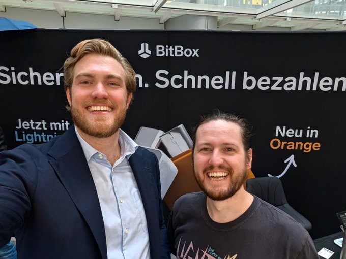 A casual selfie of two smiling young men standing in front of a 'BitBox' branded banner with German text 'Sicher... Schnell bezahlen' (Safe... Pay quickly) and an orange Bitcoin hardware wallet advertisement.