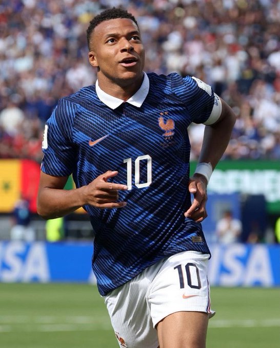 A photograph of Kylian Mbappé wearing the France national team jersey (number 10) on a football pitch, celebrating with his hand on his chest during a match.