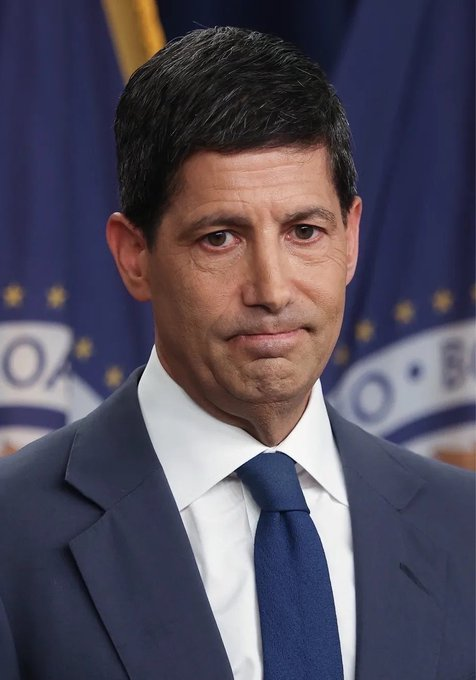 A serious-looking man with dark hair, wearing a navy suit and blue tie, stands in front of what appears to be a U.S. government seal backdrop. The image likely depicts a Federal Reserve official, relating to the news about rising odds of an interest rate hike this week.