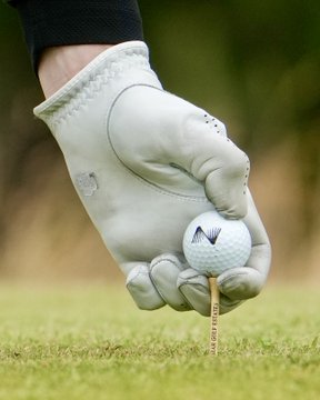 A gloved hand places a golf ball bearing the "V" Nexo logo on a tee in the grass, visually playing on the phrase "Touching grass: Nexo style."