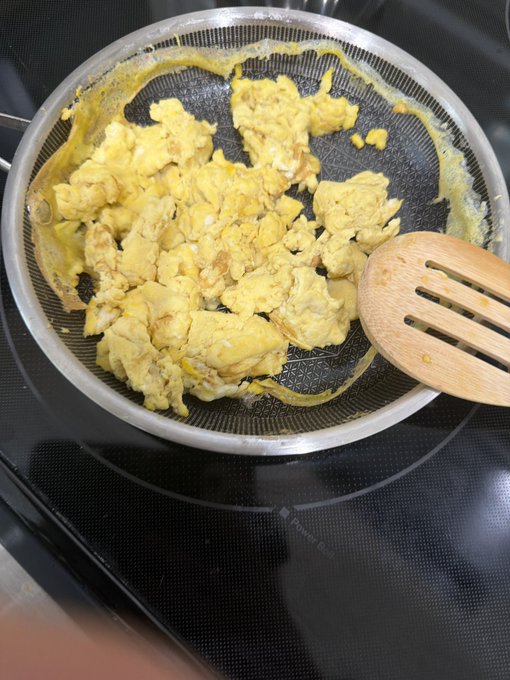 A photo of scrambled eggs cooking in a silver non-stick frying pan with a wooden slotted spatula resting on the edge, sitting on a black glass stovetop.