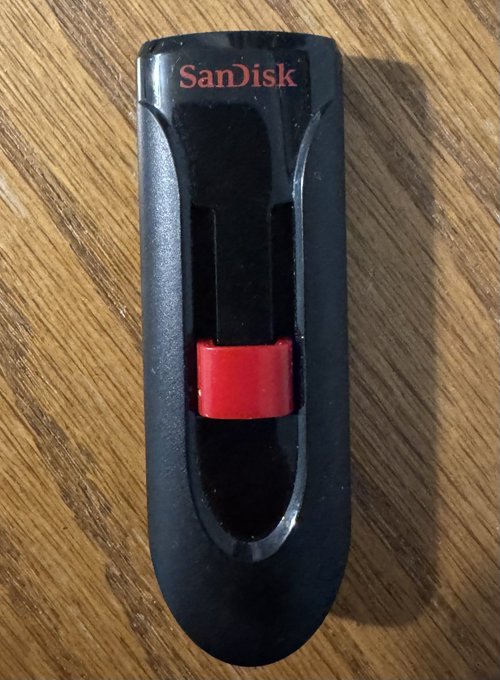 A black SanDisk USB flash drive with a red sliding mechanism, photographed on a wooden surface.