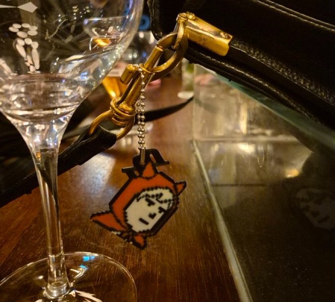 A close-up photo of a black leather handbag with gold hardware, featuring an orange charm with the Japanese text 'やって' hanging next to a wine glass.