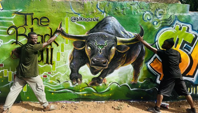 Two young men pose playfully with outstretched arms in front of a vibrant green graffiti mural featuring 'The Black Bull' painted by @bknoiz06.