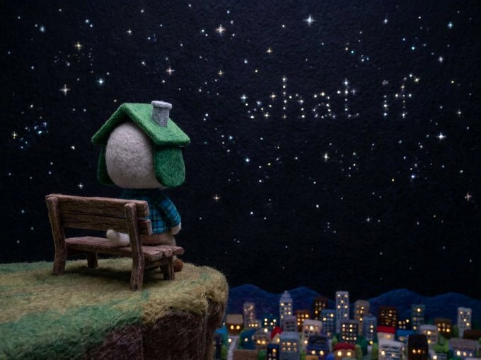 A small felt figure wearing a green hat sits on a wooden bench on a grassy cliff, gazing up at a starry night sky above a tiny illuminated town.