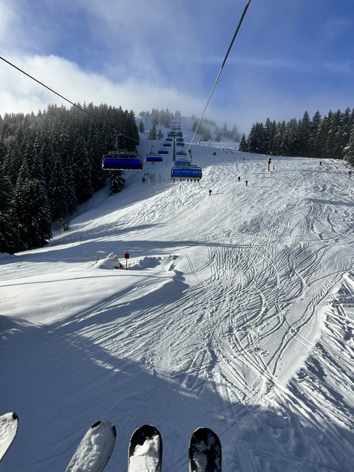 A snowboarder captures the tip of their board at the edge of a freshly carved piste beneath a ski lift, with distant skiers dotting the sunlit slope as a serene mountain backdrop. The image pairs with the caption's "gm future billionaires" mantra—visualizing the abundance and prosperity the post speaks to through the wide-open, untouched powder and limitless horizon ahead.