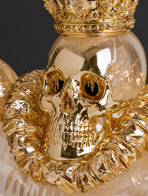 A close-up of an ornate, shiny golden skull wearing a crown, surrounded by golden ruffled fabric.
